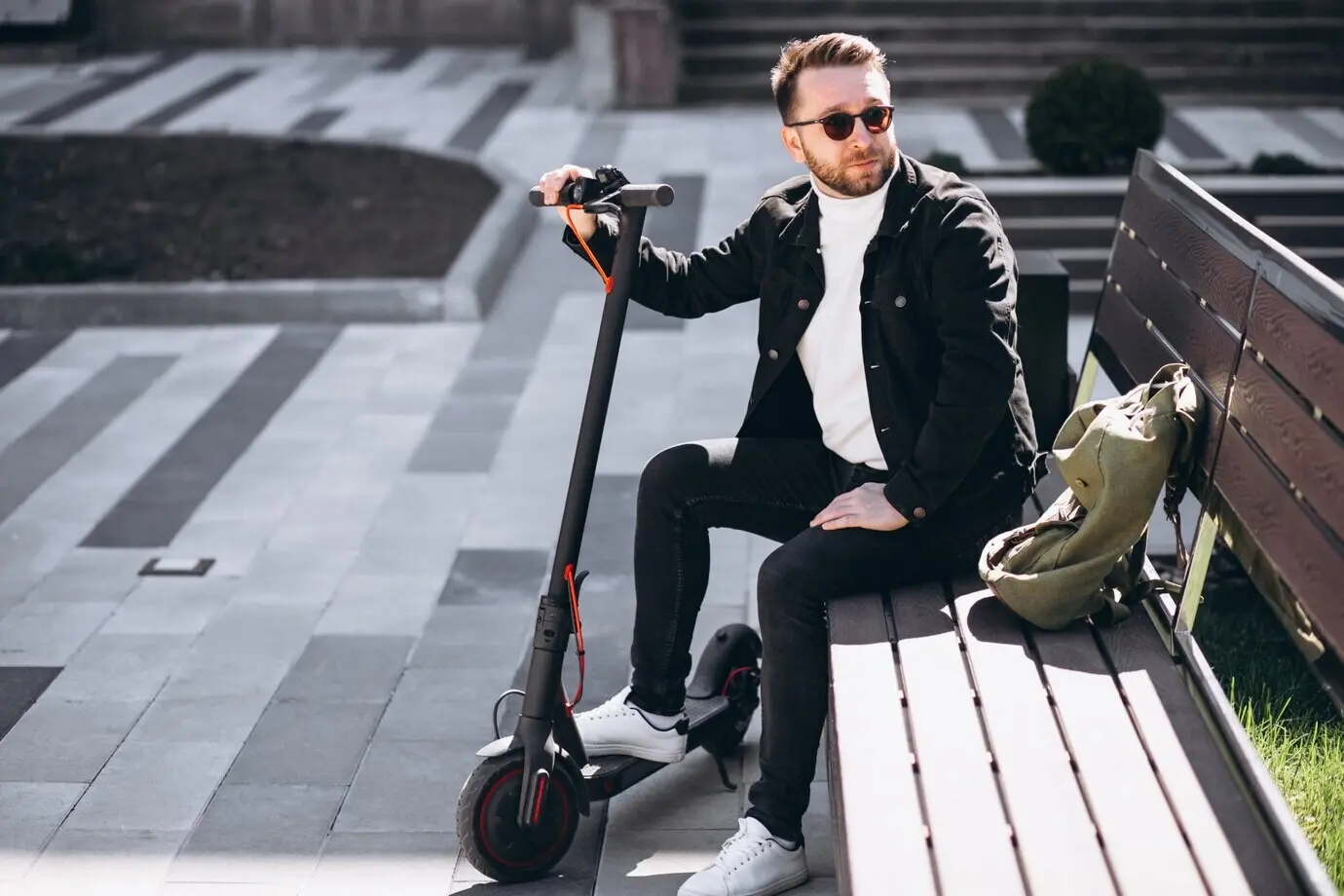 A young, handsome man sits on a park bench, taking a break from riding a scooter.